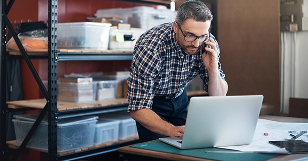 A man is  on the phone, while leaning over to look at a laptop 